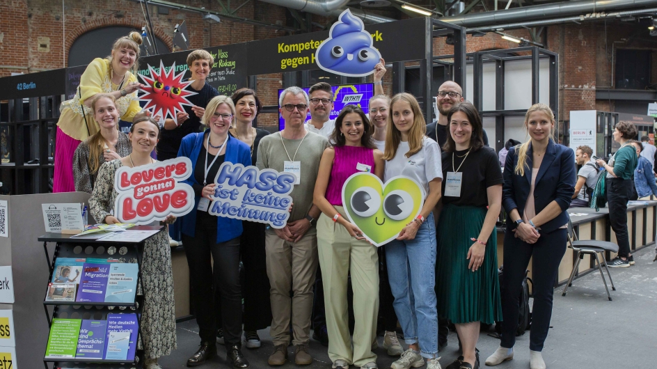 Eine Gruppe von Menschen posiert fröhlich zusammen, einige halten bunte Schilder mit Aufschriften wie „Lover's gonna love“ und „Hass ist keine Meinung“. Im Hintergrund sind Informationsstände und Displays zu sehen. Die Atmosphäre ist lebhaft und einladend.