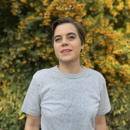 Person mit kurz geschnittenen braunen Haaren trägt ein graues T-Shirt und steht vor einem Hintergrund von grünem Gebüsch mit orangefarbenen Beeren.